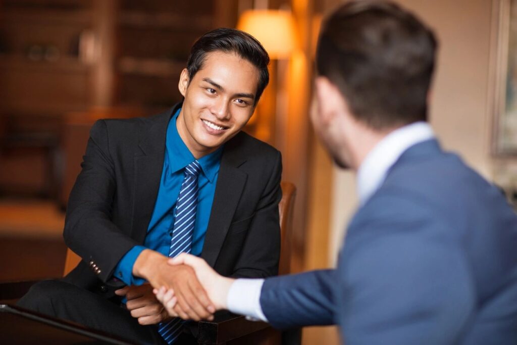 Two men Shaking each others hands, One of them is backing the camera and the other is smiling and is clearly seen.